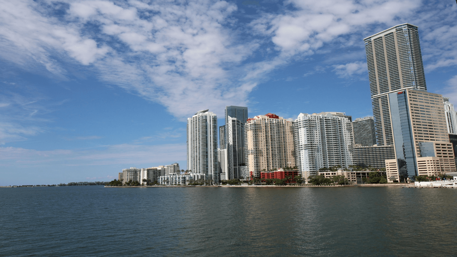 A city skyline with modern high-rise buildings along a waterfront under a partly cloudy blue sky.